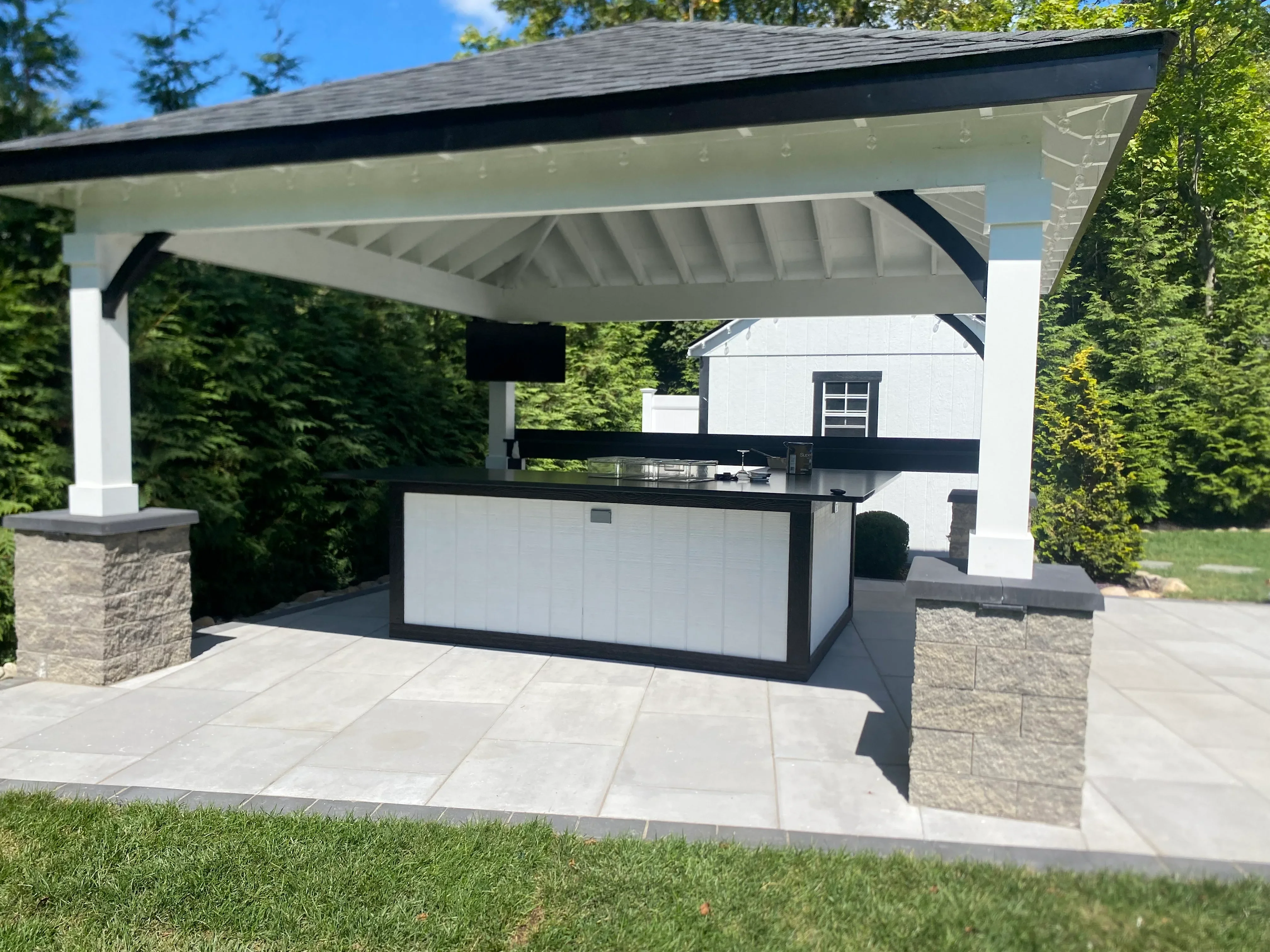 Outdoor kitchen island with white cabinetry, black trim, and stainless steel countertop under modern pergola